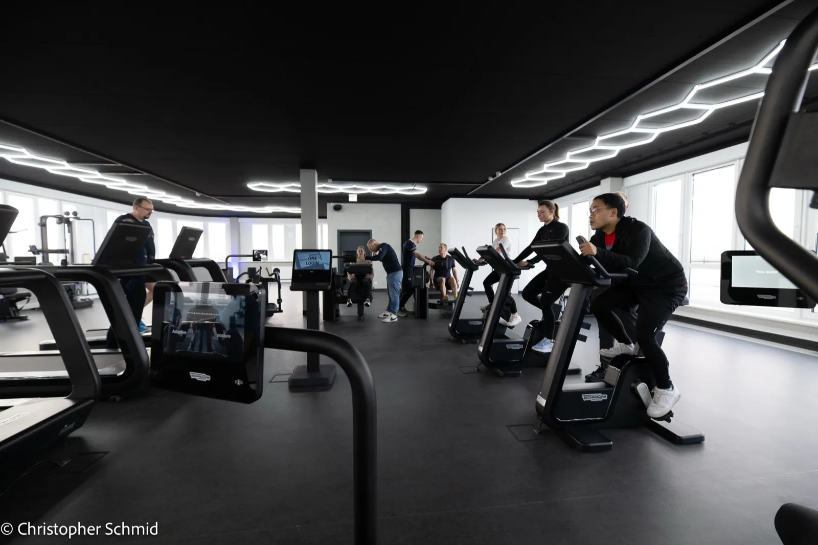 Blick über die Trainingsfläche mit Ergometern und Laufbändern unter langen Lichtbändern; mehrere Menschen trainieren.